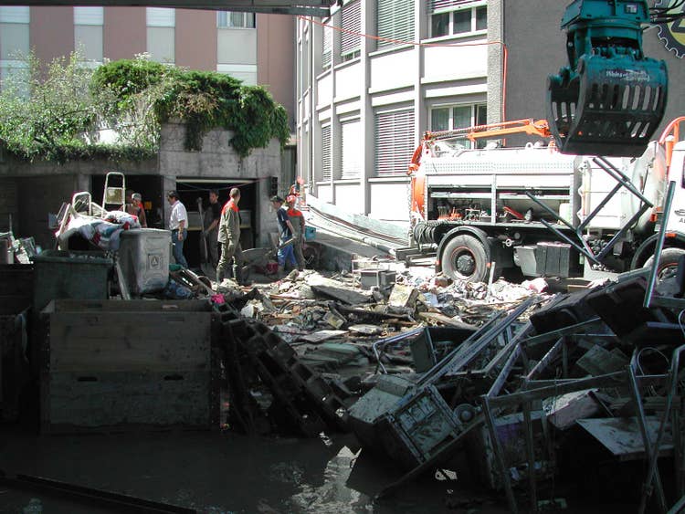 Auf dem Platz vor der Metallbauwerkstatt stapelt sich deren gesamtes Interieur. Menschen haben Schaufeln und Schubkarren in den Händen.