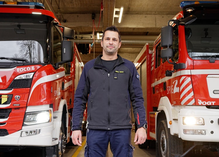 Kommandant Martin Scherz steht im Feuerwehrmagazin Spiez zwischen zwei Feuerwehrfahrzeugen und blickt direkt in die Kamera.