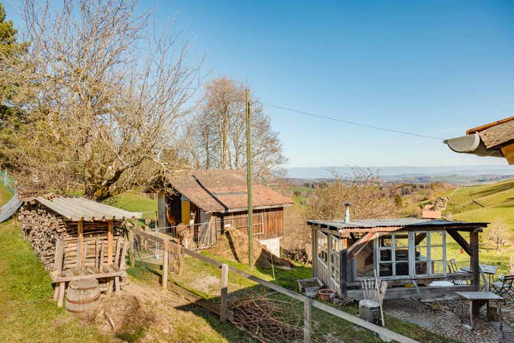 Links auf dem Bild sieht man einen Stall. Davor stehen ein paar Lamas. Rechts davon befindet sich ein Aussensitzplatz.