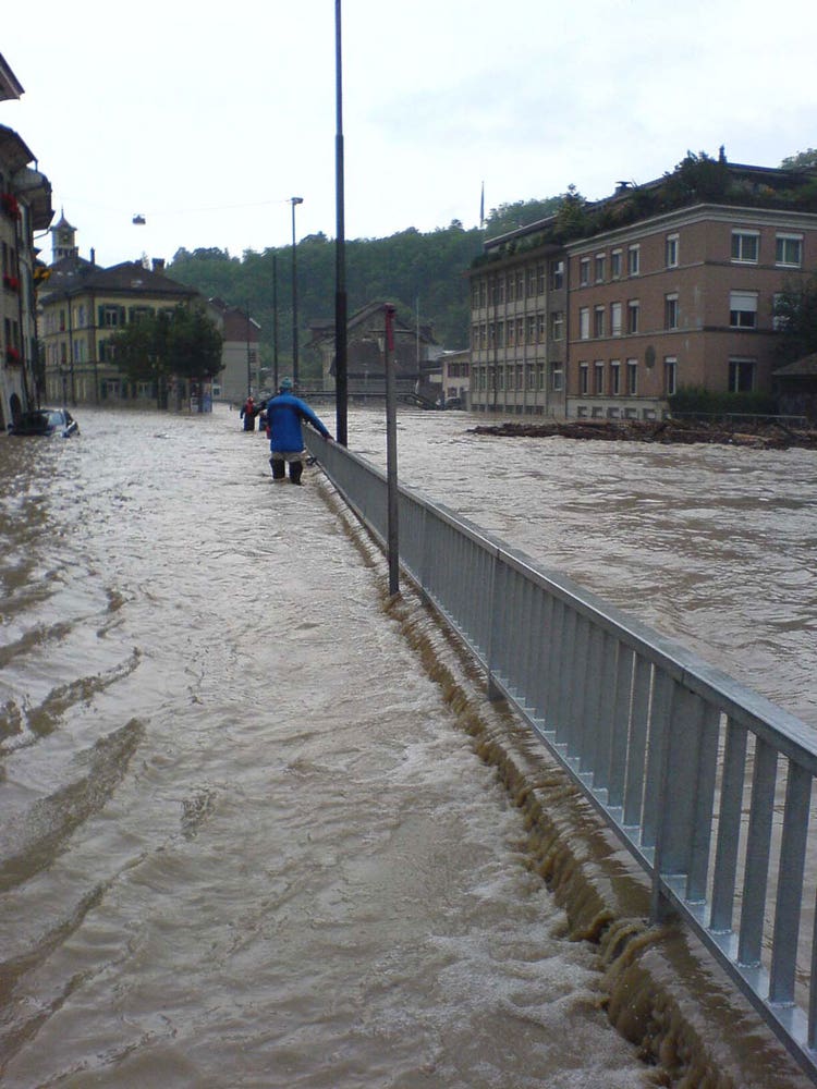Ein Mann steht bis zu den Knien im Wasser und watet an einem Geländer entlang eine überflutete Strasse hinauf.