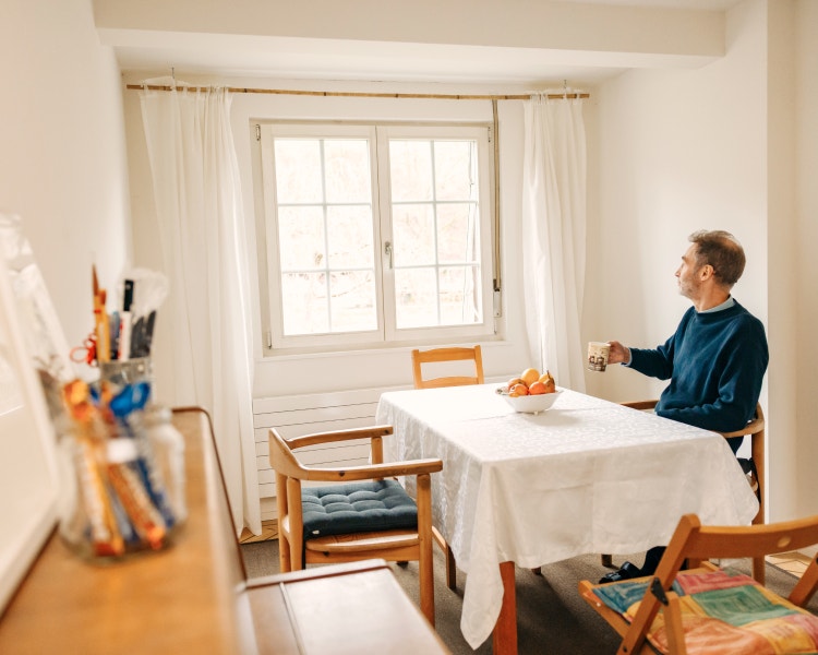 Ein Mann sitzt mit einer Tasse am Esstisch und schaut aus dem Fenster.