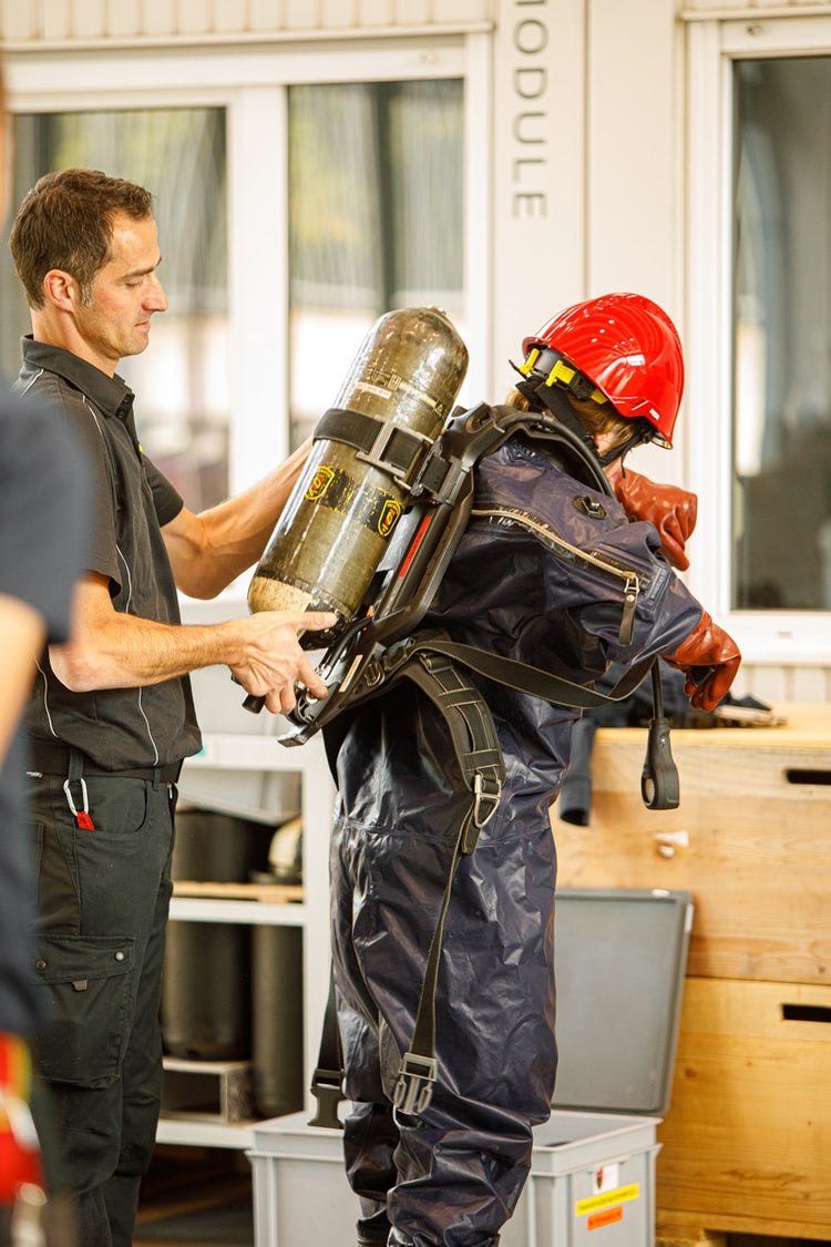 Ein Feuerwehrmann hilft einem anderen Feuerwehrmann beim Anziehen des Atemschutzgerätes.