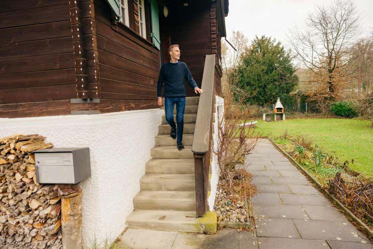 Ein Mann geht eine Aussentreppe an einem Chalet hinunter. Vor dem Haus befindet sich ein Holzstapel, im Garten ein Grill.