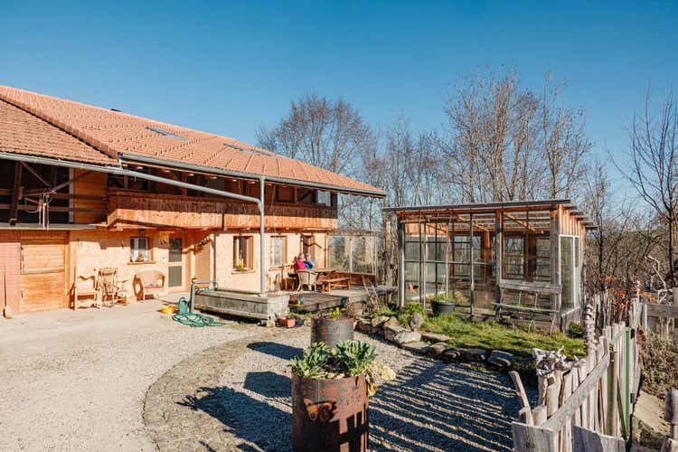 Rechts im Bild sieht man die Ostseite des Bauernhauses inklusive des Hauseingangs und einer kleinen Terrasse. Rechts davon ein Gewächshaus.