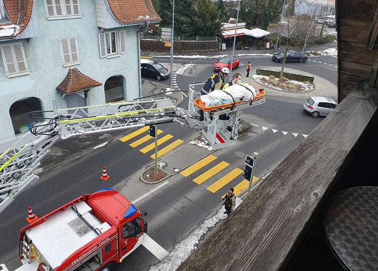 Eine Person liegt auf einer Rettungsbahre. Die Rettungsbahre befindet sich auf dem Rettungskorb einer Drehleiter eines Feuerwehrautos. Im Rettungskorb sind ein Feuerwehrmann und ein Rettungssanitäter.