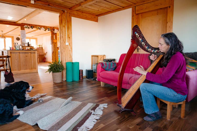 Sandra Haunreiter assise sur un petit tabouret et jouant de la harpe. La chienne Elly est allongée sur le tapis.