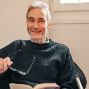 Un homme est assis dans un fauteuil avec un livre, ses lunettes de lecture à la main.