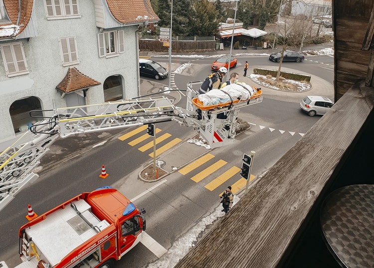 Une personne est allongée sur une civière de sauvetage. La civière de sauvetage se trouve sur la nacelle de sauvetage de la grande échelle d’un camion de pompiers. Dans la nacelle de sauvetage se trouvent un pompier et un secouriste.