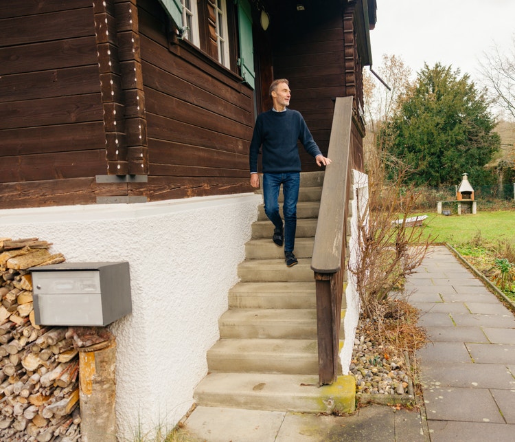 Un homme descend l’escalier extérieur d’un chalet. Devant la maison, il y a une pile de bois, et dans le jardin se trouve un barbecue.