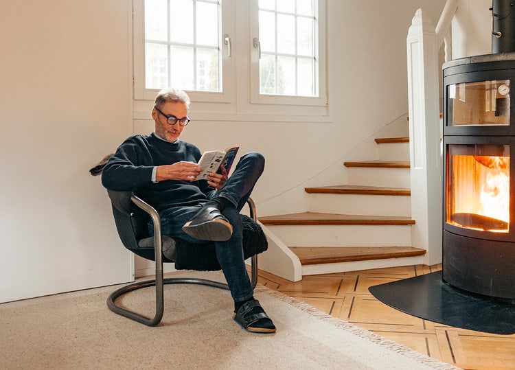 Un homme est assis dans un fauteuil de lecture avec un livre. À côté de lui, un feu brûle dans un poêle suédois.