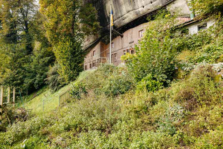 Le Fluehüsli, construit dans les rochers de grès, entouré d’une végétation dense, en haut d’un talus abrupt. La roche en saillie coiffe le toit.